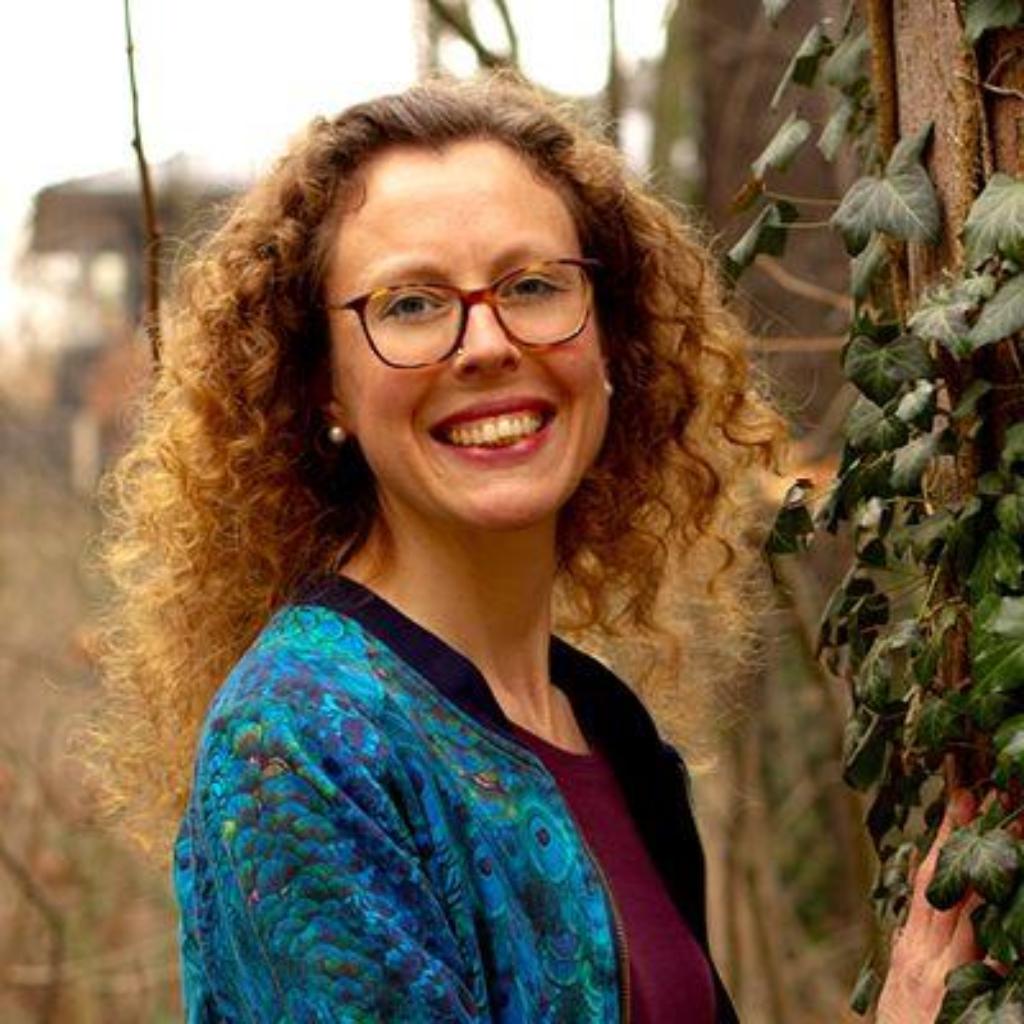 Porträt von Maria Giering: Lächelnde Frau mit lockigem Haar und Brille im Freien neben Efeu, trägt eine blau gemusterte Jacke